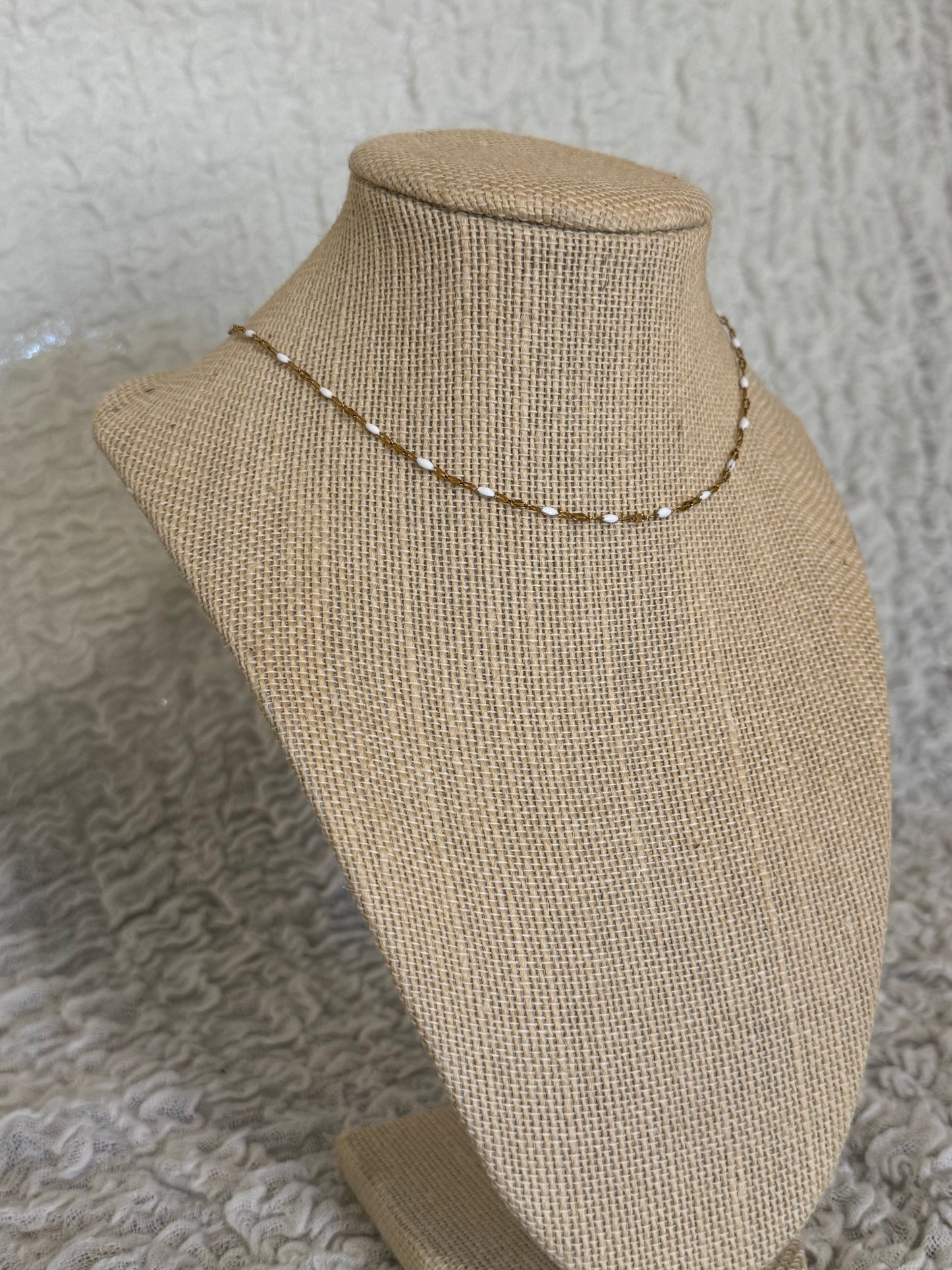 A delicate gold beaded necklace displayed on a beige burlap jewelry bust against a textured gray background.