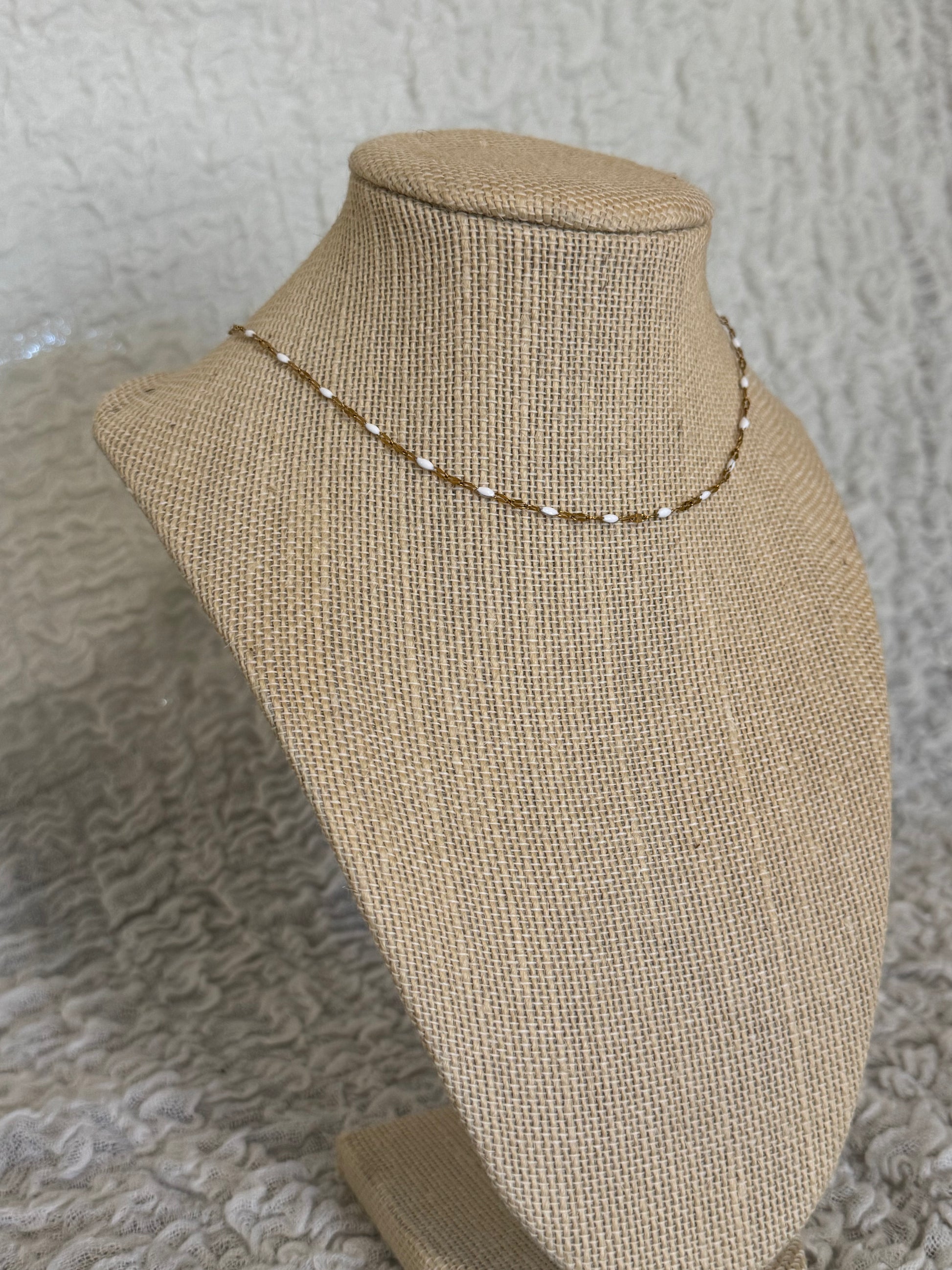 A delicate gold beaded necklace displayed on a beige burlap jewelry bust against a textured gray background.