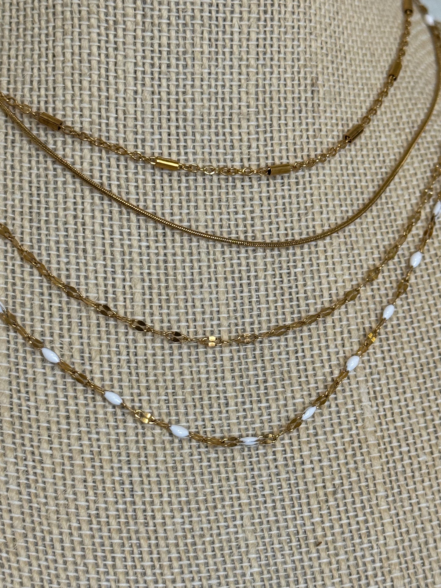 A layered necklace set displayed on a burlap-textured mannequin bust. The set includes a gold chain, a gold chain with small dark metallic beads, a gold chain with white oval beads, and a gold chain with tiny gold and white beads.