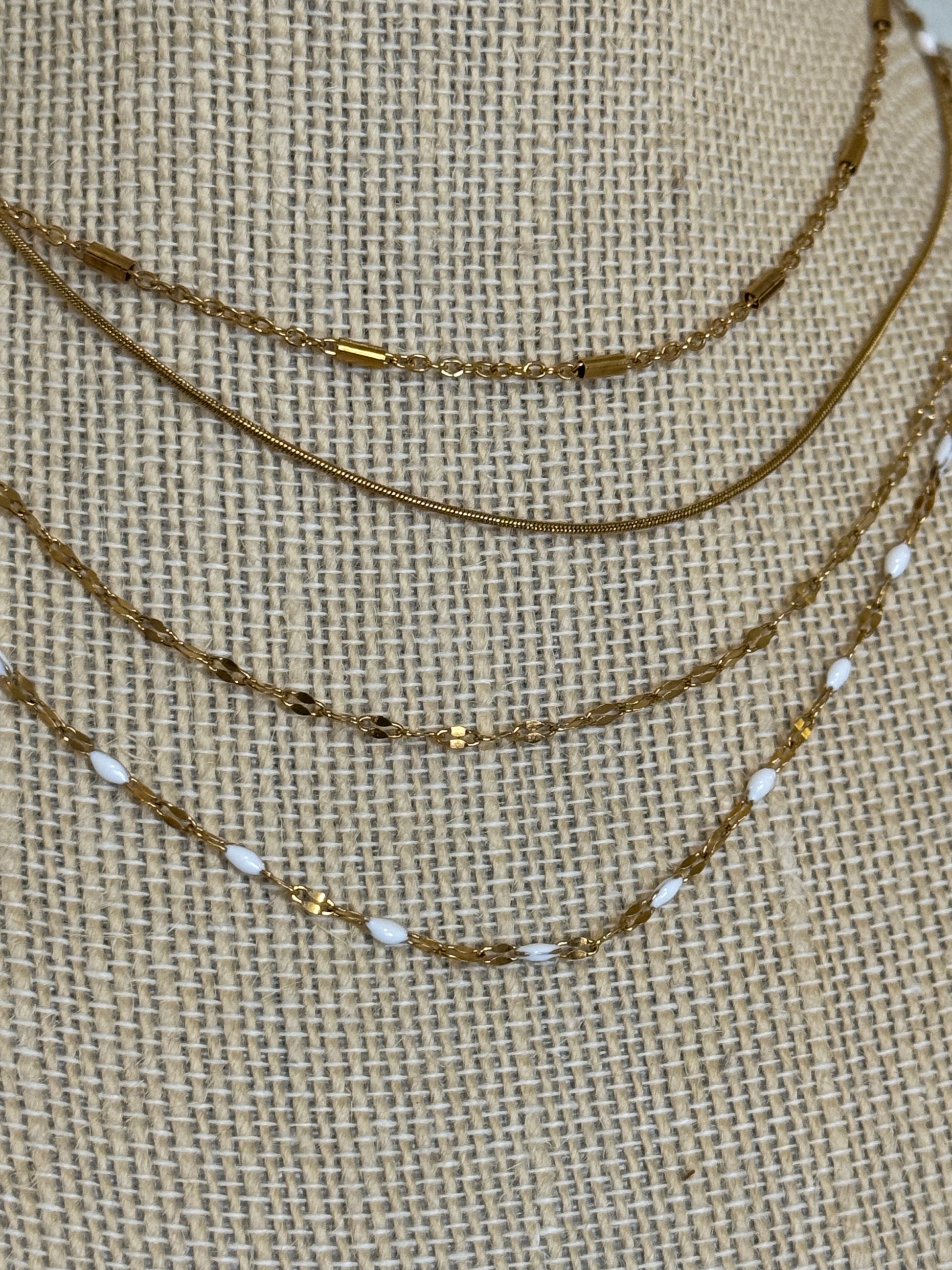 A layered necklace set displayed on a burlap-textured mannequin bust. The set includes a gold chain, a gold chain with small dark metallic beads, a gold chain with white oval beads, and a gold chain with tiny gold and white beads.